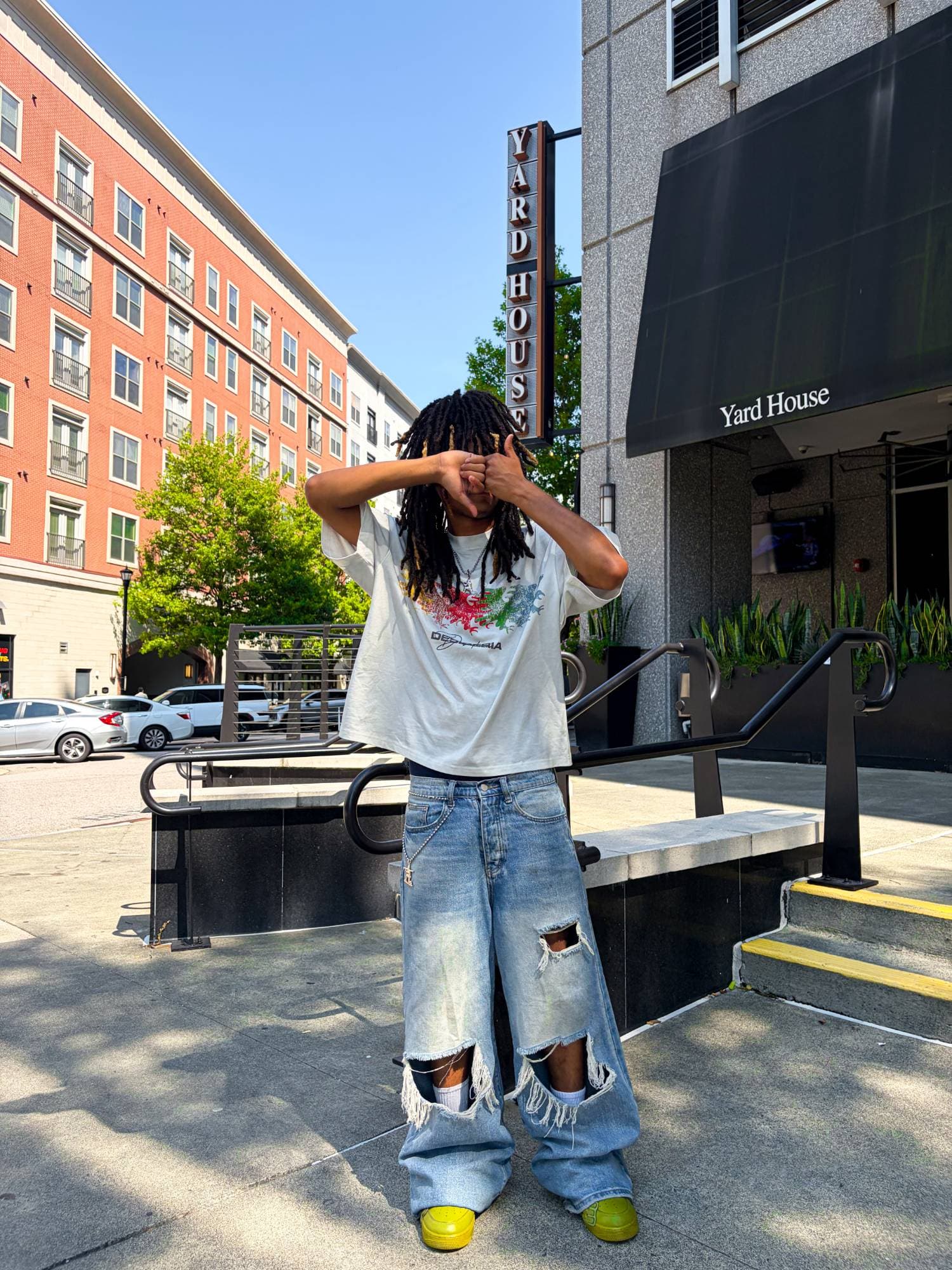 Forearms crossed over the face outside Yard House, baggy ripped light-wash denim